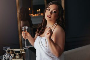 Woman wrapped in a white towel beside a clawfoot bathtub holding a champagne glass during a glamorous boudoir photography session by Black Lace Boudoir.