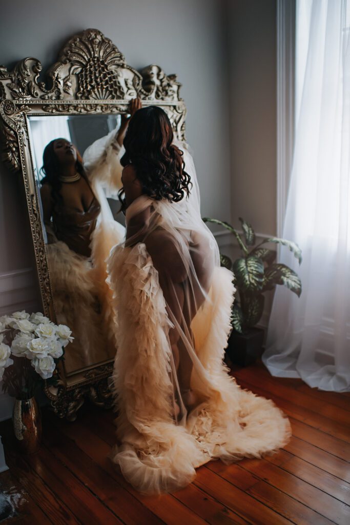 Ms. B stands in front of a large, gold framed mirror. She wears a luxury beige inheritance robe and a faux leather bodysuit for her birthday boudoir session. 