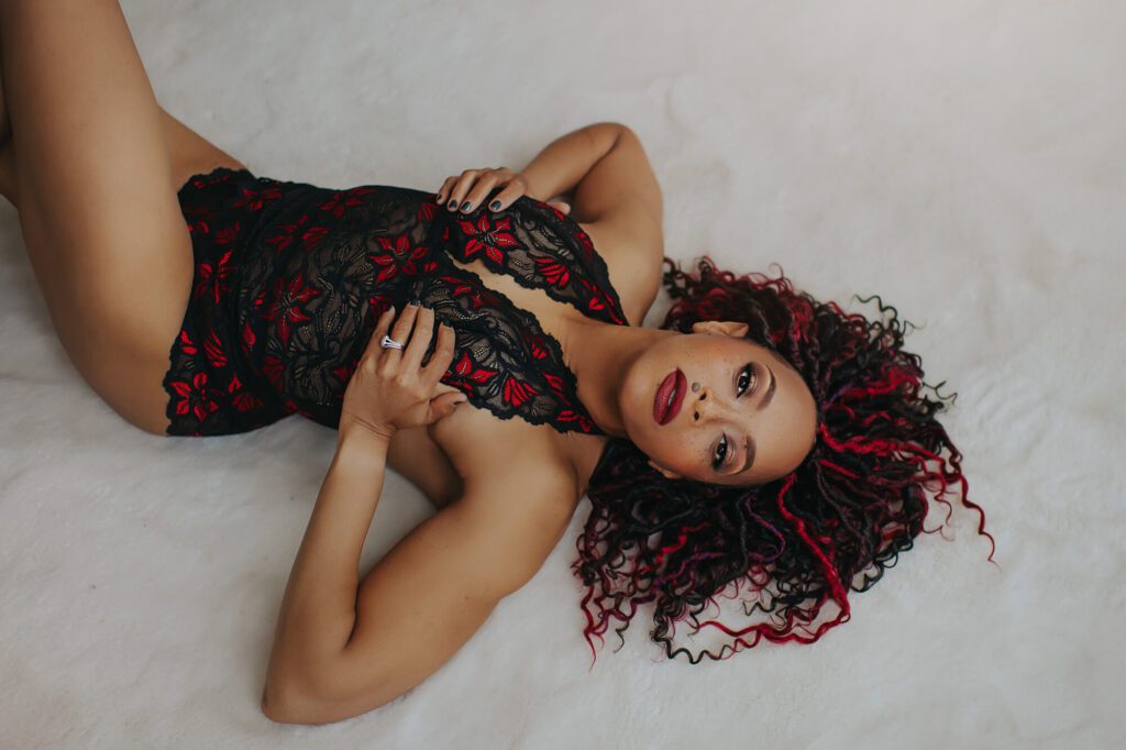 Woman reclining on a soft white backdrop in red and black lace lingerie during a luxury boudoir photography session by Black Lace Boudoir.